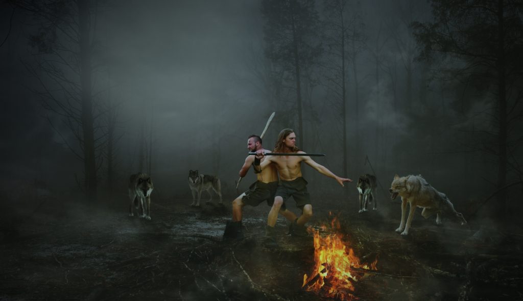two light-skinned men wearing shorts and low boots, holding longswords, standing back-to-back, surrounded by 4 wolves. A campfire burns in the foreground.