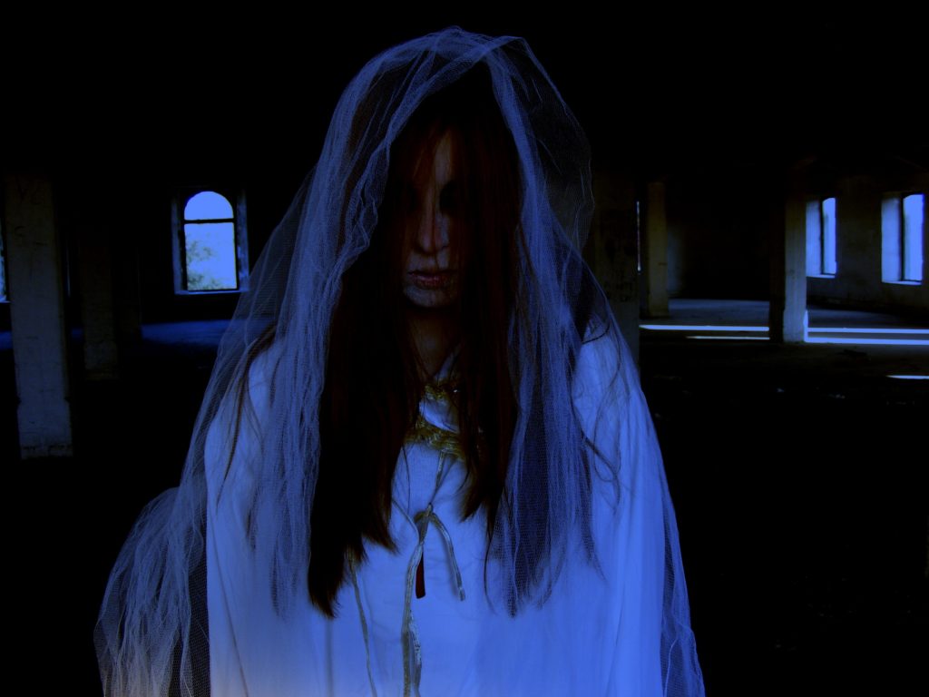 In a dark building with rows of benches and light coming in a few arched windows, a white woman with darkened eyes and dark red-brown hair covering most of her face.