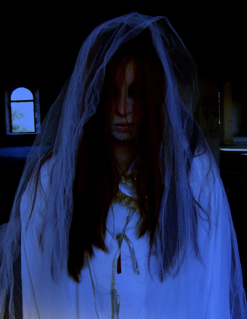 In a dark building with rows of benches and light coming in a few arched windows, a white woman with darkened eyes and dark red-brown hair covering most of her face.