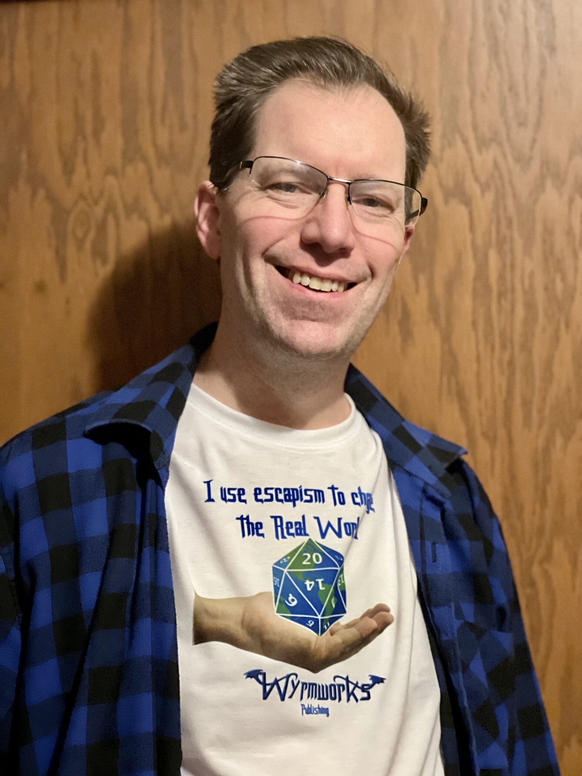 Dale Critchley, a middle-aged white man with short blond hair, wearing a blue plaid flannel shirt with a white t-shirt beneath with a hand holding a D20 globe. Above the globe, "I user escapism to change the real world," and below, the Wyrmworks Publishing logo, standing if front of a wooden door