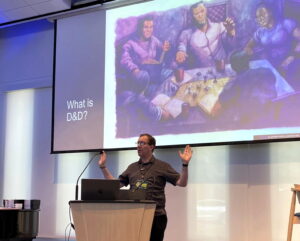 Dale, a middle-aged white man, standing at a podium, talking with hands in the air, a screen behind him showing players at a D&D table and What Is D&D?